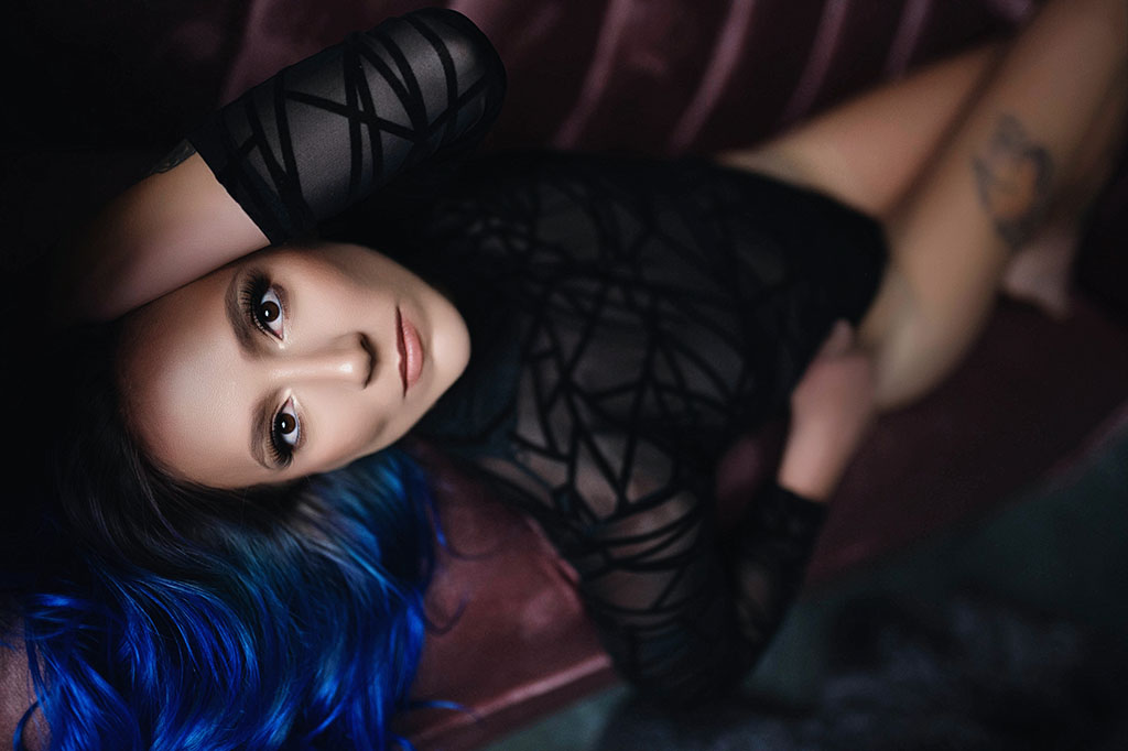 women with beautiful blue hair laying on a pink couch in a black bodysuit for her boudoir photo shoot.